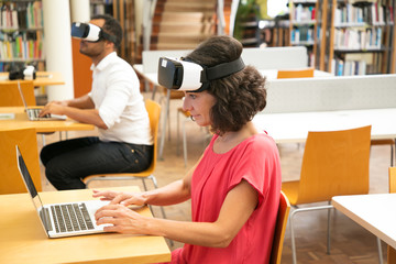 Group of students using VR experience for work on research. Middle aged woman in VR headset sitting at desk and working on laptop. Students with VR glasses concept