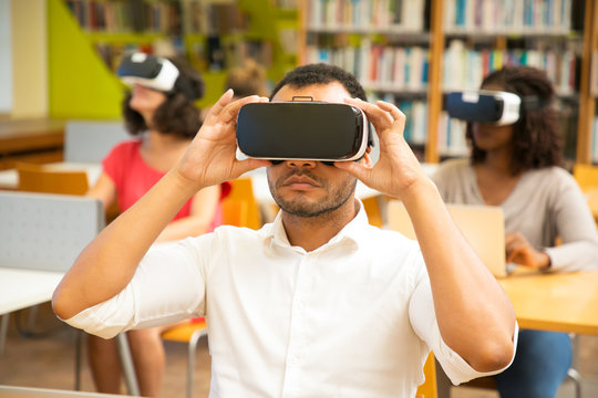 Mix raced group of students watching virtual video tutorial in library. Man and women wearing virtual reality headsets, sitting at desks with laptops. VR video concept