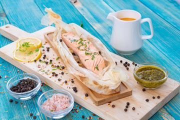 Baked Salmon in Grilled Paper with Lemon Slices and Spices (Salt and Pepper) on a Wooden Plate with Sauce with some Greens on Top. Wood Blue Background