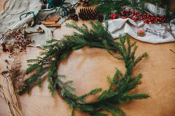 Fototapeta na wymiar Fir branches, pine cones, thread, berries, cinnamon on wooden table. Rustic Christmas wreath workshop. Authentic stylish still life. Making Christmas wreath.