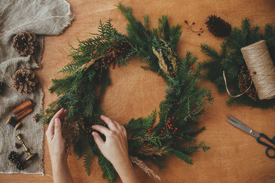 Hands holding christmas wreath with fir branches, berries, pine cones, and thread, scissors on rural wooden table. Rustic Christmas wreath, flat lay. Christmas wreath workshop