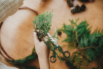 Making rustic christmas wreath. Hands holding cedar branches and pine cones, thread, scissors on wooden table. Details for workshop of making christmas wreath