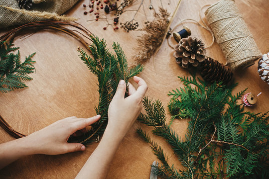Hands holding fir branches and pine cones, thread, scissors on wooden table. Details for workshop of making christmas wreath. Making rustic christmas wreath