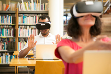 Students using VR simulators for studying. Man and woman in virtual reality headsets sitting at desks with laptops and touching air. VR technology for education concept