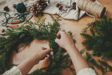 Hands holding fir branches, and pine cones, thread, berries, scissors on wooden table. Christmas wreath workshop. Authentic stylish still life. Making rustic Christmas wreath. - obrazy, fototapety, plakaty