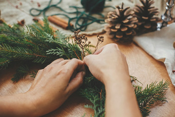 Hands holding herbs and  fir branches, pine cones, thread, berries, cinnamon on wooden table. Christmas wreath workshop. Authentic stylish still life. Making rustic Christmas wreath.