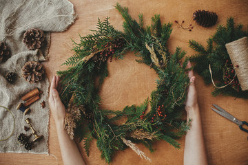 Rustic Christmas wreath, flat lay. Hands holding christmas wreath with fir branches, berries, pine cones, and thread, scissors on rural wooden table. Christmas wreath workshop