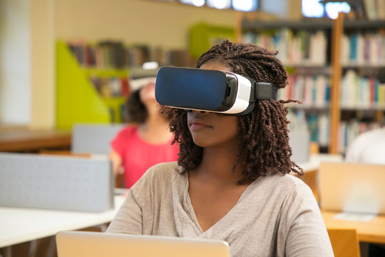Interracial group of students using VR experience for work in library. Young black woman wearing virtual reality headset, sitting at desks, using laptop. Innovation technology concept