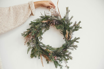 Hipster girl in white sweater holding rural christmas wreath with fir branches, berries, pine cones and herbs  on white wall in room. Christmas rustic wreath. Season's greetings © sonyachny
