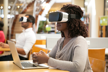 Adult students using VR simulators for work on project in library. Man and woman wearing virtual reality glasses, sitting at desks with laptops, holding air. Augmented reality concept - Powered by Adobe