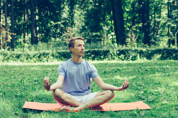 Obraz premium Young attractive man in sport clothes is meditating in the lotus position with a pacified face in the park against the background of green grass and trees.