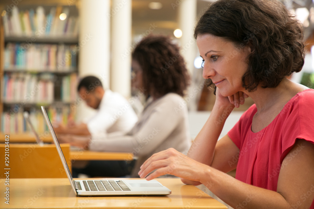Wall mural satisfied female user using wireless internet connection in public library. smiling middle aged woma