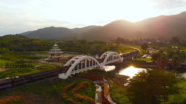 Aerial view of Thailand express train runs pass through the old white bridge during sunset or sunrise with flare light. Old white bridge a tourist attraction famous of Northern Thailand.