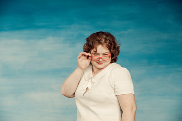 Attractive red-haired plump woman in a white suit adjusts red glasses on a blue  background, looks in surprise at the frame.