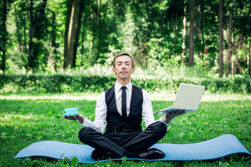 Business man relaxing in a park in the lotus position, he can't relax in any way,  in one hand...