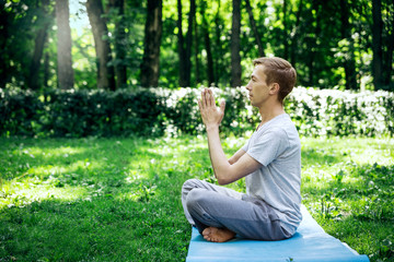 Naklejka na ściany i meble A young attractive man practices yoga in nature and meditates with his eyes closed in the lotus position on a green lawn.