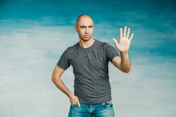 A young bald man in a gray T-shirt on a blue background surprised looking at the camera, raising his palm 