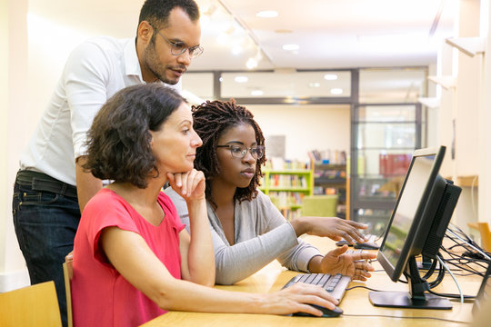 Tutor helping students in computer class. Man and women sitting and standing at desk, using desktop, pointing at monitor and talking. Education concept