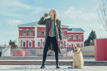 A handsome young guy in a khaki and berets military coat walks with a big dog. Drinks from flask in...
