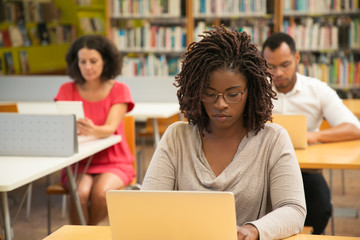 Serious African American student studying in library computer class. People sitting at desks and using laptops in classroom with bookshelves. Studying concept