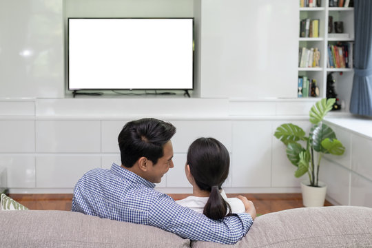Back view of young family, man and woman watching TV together in living room, TV on white background