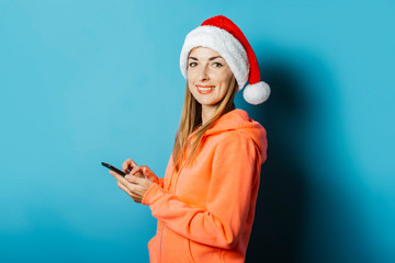 Beautiful Young woman with a smile in a hoodie and a hat of Santa Claus holds a mobile phone on a blue background. Concept winter, christmas, winter holidays, application