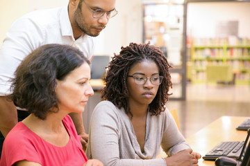 Adult female students consulting instructor in computer class. Man and women sitting and standing at desk, using desktop, looking at monitor, talking. Training concept