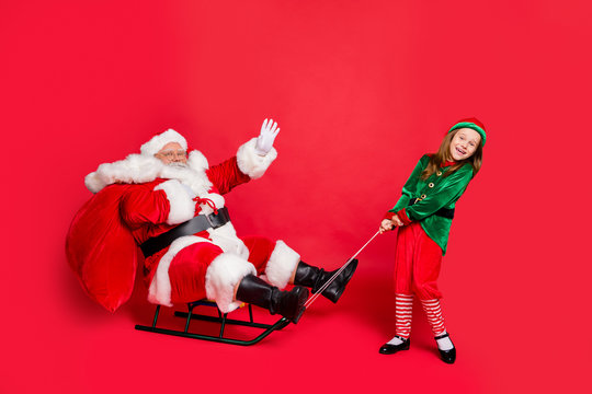 Full length photo of lovely two fairy santa claus elf with redhair grey hair waving pulling sledge carrying bag wearing green hat headwear eyewear eyeglasses isolated over red background