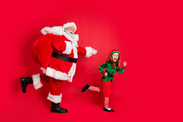 Full body profile side photo of cheerful two fairy runners santa claus elf in hat headwear carrying heavy bag with presents on midnight wearing eyeglasses eyewear spectacles isolated over red