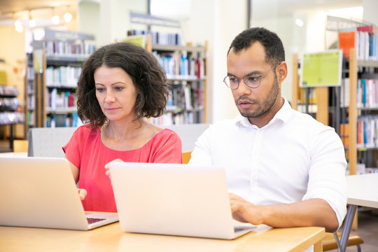 Adult Student Cheating During Test In Library Computer Class. Man In Casual Sitting At Desk, Using Laptops, Looking At Monitor Of College Mate. Exam And Cheating Concept