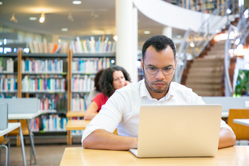 Focused male adult student doing research in library. Serious Latin man sitting at desk and using laptop. Bookshelves in background. Academic research concept