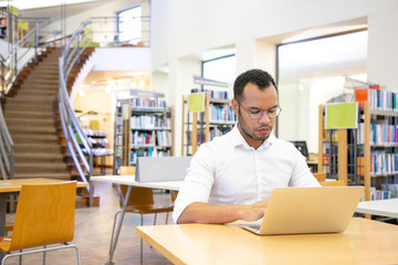 Serious adult student doing research in library. Latin man in formal shirt and eyeglasses sitting at desk, using laptop, typing. Bookshelves in background. Communication concept