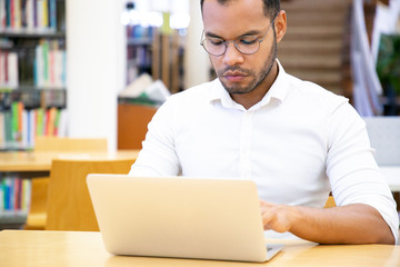 Serious adult student working on project in library. Latin man in formal shirt and eyeglasses sitting at desk, using laptop, typing. Bookshelves in background. Learning concept