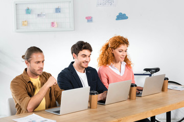 three smiling friends sitting at table and using laptops in office