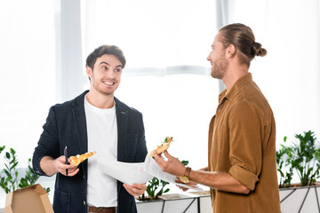 two smiling friends holding pizza slices and papers in office