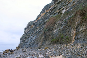 Rainy weather on sea coast with stone beach near huge rocks. Seascape in overcast day. White foggy sky over blue ocean water and mountain cliff. Outdoors nature background.