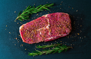 Close-up the fresh raw meat, beef steaks on the dark slate board with pepper and rosemary for the healthy diet food cooking preparation concept