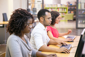 Multiethnic group of students studying in computer class. Line of man and women in casual sitting at table, using desktops, typing, looking at monitor. Training course concept
