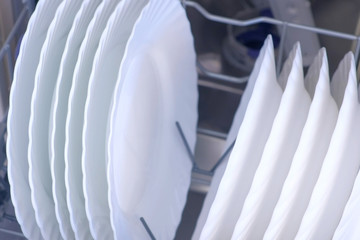 Clean white dishes and plates in basket of built-in dishwasher in kitchen. Open dishwasher mashine with tableware, closeup view.