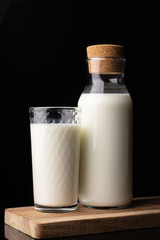 A glass filled with milk and a bottle with milk on a wooden board. Black background. Glass bottle. Natural cork. White drink. Cow's milk.