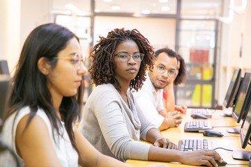 Diverse group of trainees working in computer class. Line of man and women in casual sitting at table, using desktops, looking at monitor or camera. Training concept