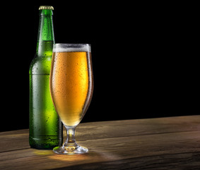 Glass of light beer and green beer bottle on the bar counter on a black.