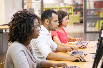 Diverse group of adult students working in computer class. Line of man and women in casual sitting at table, using desktops, typing, looking at monitor. Training center concept