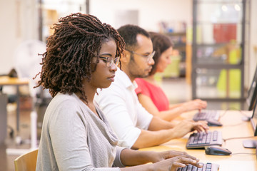 Diverse group of trainees studying in computer class. Line of man and women in casual sitting at table, using desktops, typing, looking at monitor. Computer class concept