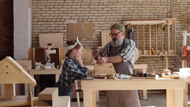Medium shot of Caucasian girl leaning on workbench, watching attentively good-looking senior craftsman screwing screws in box with electrical screwdriver