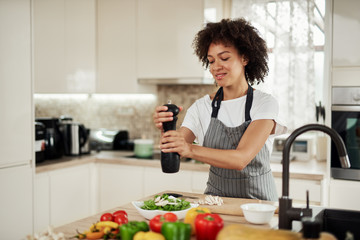 Attractive mixed race woman in gray apron addinf pepper in bowl with rocket and mushrooms. On kitchen counter are different vegetablet.