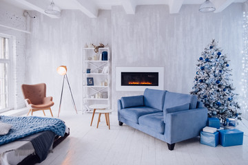 New Year's interior of the apartment. Big Windows, blue sofa, artificial fireplace, bed, lights, white and blue Christmas tree, armchair and stylish floor lamp. Loft style. Concrete wall. Cozy house.