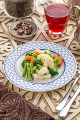 Mixed vegetables cauliflower, broccoli, bean and carrots on white plate and glass of red drink on oriental wooden table