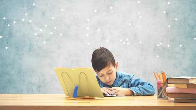 Young concentrated school kid in blue jeans t-shirt writing, copying the text from book on the bookholder.