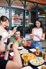 Group of Asian young people making photos of meal together on their mobile phones while they have a dinner at the restaurant outdoors